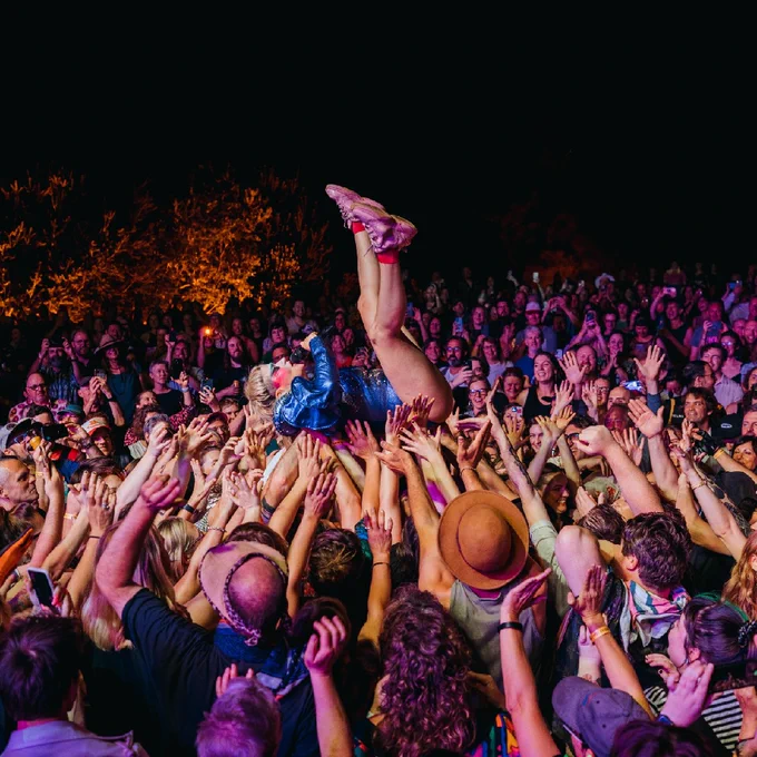 A crowd of people are holding the singer Peaches in the air, she is laying on her back with her legs kicked high. She wears a metallic blue jacket.