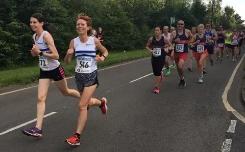 A group of runners in a road race. Two women in the lead. One is turned to the camera and smiling.