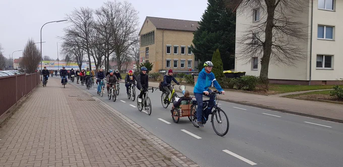 Eine Gruppe Menschen auf Fahrrädern, die eine Straße entlang fahren. Im Vordersten sitzt eine Person im Anhänger und brüllt Sprüche!