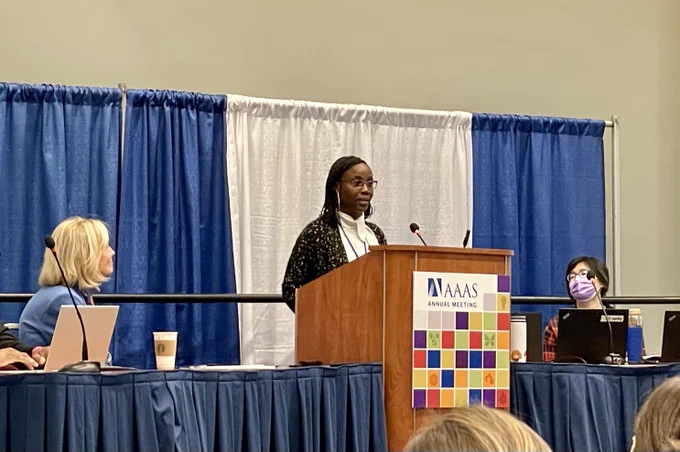 A speaker stands at a podium in front of a blue curtain with a panelist on either side.