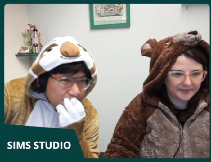 GrimSuruDoi wearing a sloth onesie staring intently at the screen with his head in his 3 toed hand while SimGuruMorgan (wearing a bear onesie) shows off the new Growing Together pack