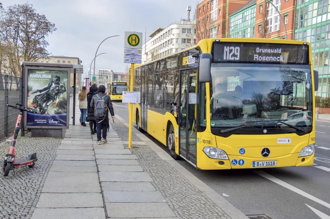 A bright yellow passenger bus waits at a stop while passengers disembark on a street in Berlin.