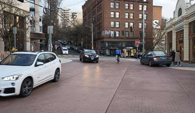 A large expanse of reddish brick with several cars and in the distance a person on a bike. Dense buildings with at-grade light shops. But no  people visible on the streets.

Same street at Pike and Melrose as previous. This photo taken by Ryan Packer, who says, What a failure this "raised crosswalk" at Pike and Melrose is. There's not really another word for it.