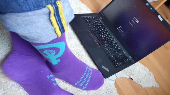 Zwei Füße mit lila Socken stehen auf dem Boden vor einem schwarzen ThinkPad-Laptop. Auf den Socken steht in türkiser Schrift "Feministisch. Solidarisch. Kämpferisch." Auf Höhe der Knöchel ist ein türkises Frauensymbol mit einer Faust in der Mitte.