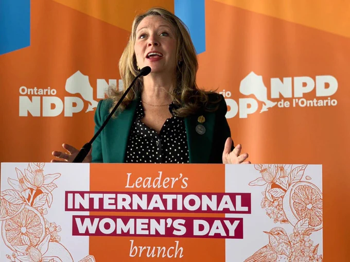 I’m standing at a lecturn. A sign on the front says “Leader’s International Women’s Day brunch.” The artwork has beautiful line illustrations of oranges, flowers, and birds.