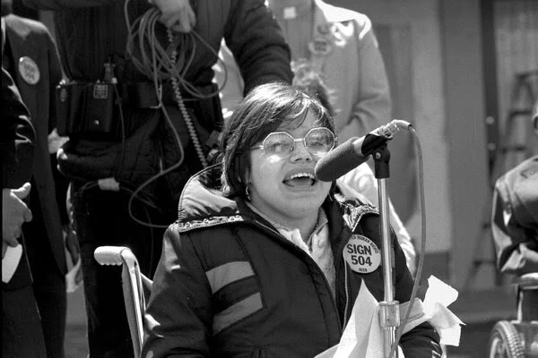 Black and white photo of a young Judy Heumann speaking at a microphone outside in her wheelchair, wearing a "Sign 504" button. 
