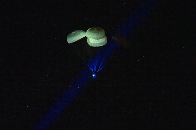 The SpaceX Dragon Endurance spacecraft is seen as it lands off the coast of Tampa, Florida on Saturday, March 11, 2023. Dragon's four white parachutes are the most dominant feature of this photo, with thin cords connecting them to the capsule below—which is lit brightly by a blue spotlight coming up from the ground. The background is black, with scattered white dots. Credit: NASA/Keegan Barber
