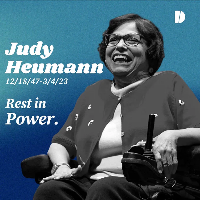 a navy and teal gradient background with white text has a black and white picture of Judy Heumann taking up the majority of the right half; she is an older woman in a power wheelchair smiling with her mouth wide. To the left is white text “Judy Heumann 12/18/47-3/4/23 Rest in Power.”  A white Diversability symbol sits in the upper right corner.