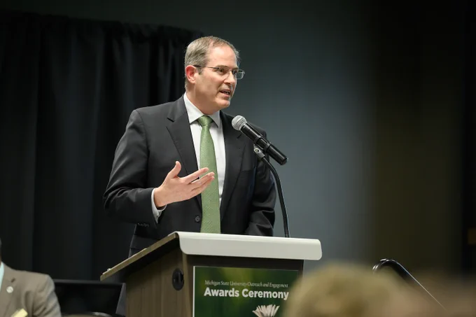 Chris Long at a podium that reads Michigan State University Outrearch and Engagement Awards Ceremony. He is looking slightly to his left, right hand raised, speaking. He is in a dark suit, grey shirt, and green tie.