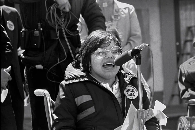 Image description: A black and white photo of Judy Heumann. In the photo, she has short, chin-length hair and octagon-shaped metal glasses and is speaking into a microphone while seated in a manual push wheelchair. She is wearing a coat with a large button pin with words that read “SIGN 504” in very large block letters and “HUMAN RIGHTS ACCD” in smaller block letters.