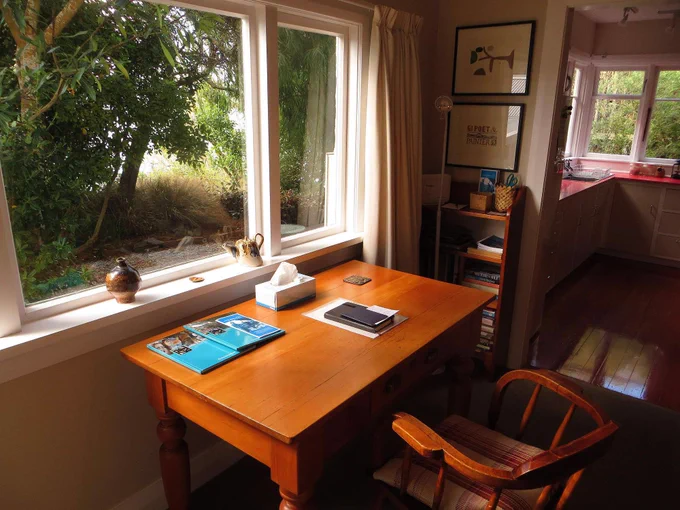 The writing space in the Caselberg House in New Zealand. There’s a wooden writing desk and chair facing a window looking out onto trees. There are some papers on the desk and a small shelf to the right with books on it.