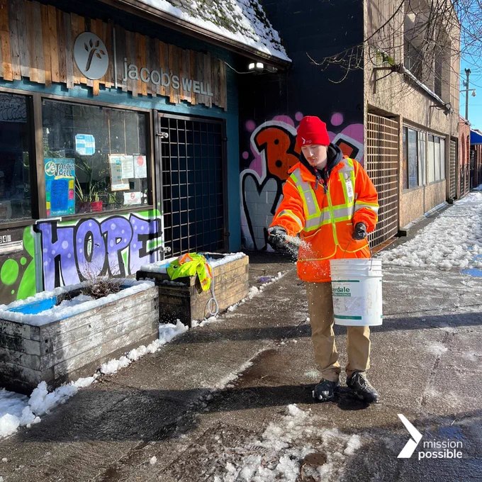 A Mission Possible associate is walking and sprinkling salt along a sidewalk while holding a large white bucket filled with salt. They are wearing an orange safety jacket, a red toque, black gloves, khaki pants, and black boots. There is a white Mission Possible logo edited on to the photo in the bottom right corner.