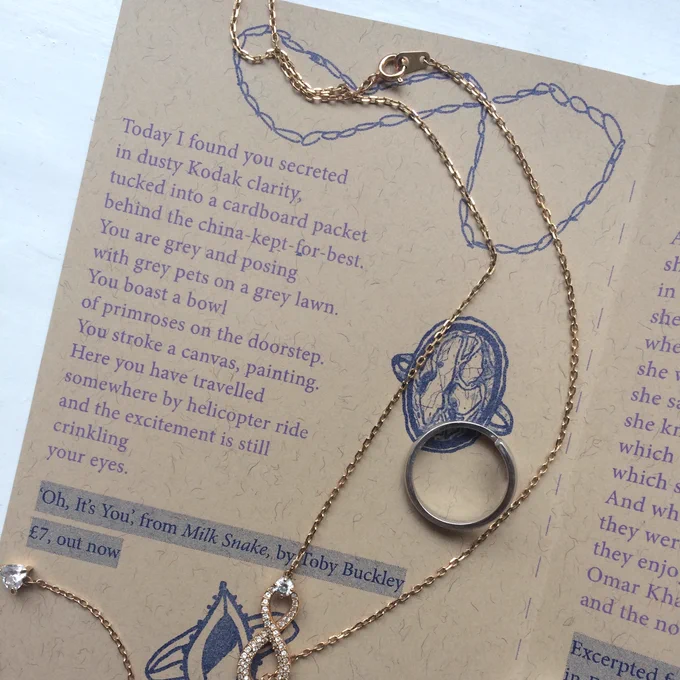 An EP Zine open on 'Oh, It's You' by Toby Buckley. A necklace snakes across the paper next to a single silver ring. Both sit on top of an illustrated chain and ring