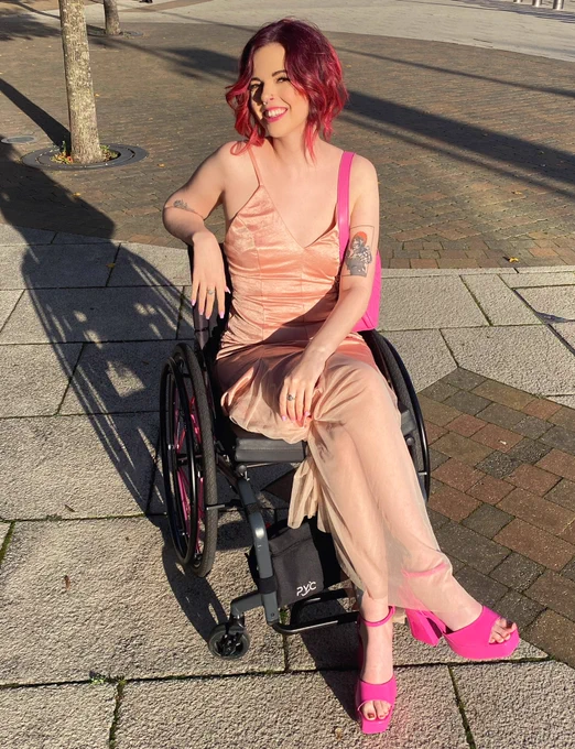 ru, a white femme person in their wheelchair. she has dark pink hair in a low updo. they are wearing a nude, full length gown and hot pink shoes with a matching bag. she is smiling at the camera with her legs crossed.