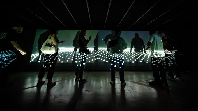 People in silhouette dance in a field of lasers against a projected backdrop, with light reflecting on the black floor.