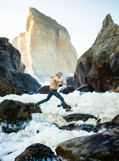 Ben Moon leaps from one rock to another, camera in hand, on the rocky shore of Oregon.