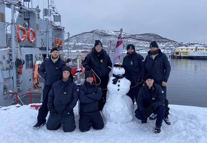HMS SMITER’s ship’s company stand around the snowman they have built with their ship in the background.