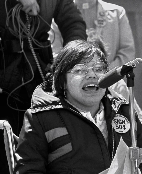 Black and white photo of Judy Heumann. In the photo, she has short, chin-length hair and octagon-shaped metal eyeglasses and is speaking into the microphone while seated on a wheelchair. She’s wearing a coat with a large button pin with texts that read “SIGN 504” in a large block letters and “Human rights ACCD” in small block letters.