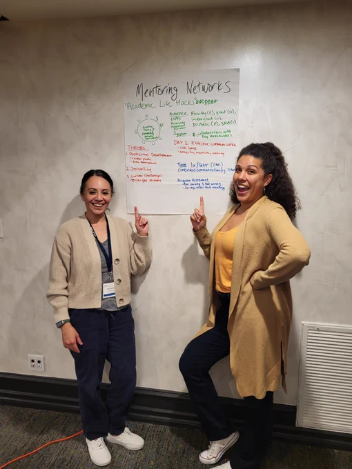 Pearl and Kacie smiling, standing in front of a wall with a poster on mentoring networks. 