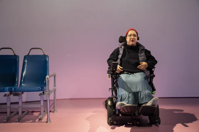 Alejandra, a light-skinned Latina with short dark hair that is slicked back, sits in her motorized wheelchair. She wears a red headband, glasses, a black sweater, and a navy-blue puffer vest. She also wears a blue denim skirt and grey fuzzy slippers. Her hands rest on her sweater. The floor beneath her is light pink with a white/pink wall behind her. Two blue bus seats with silver metal are next to her. 