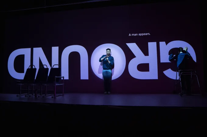 The word “GROUND” is projected upside-down in large, white capital letters. In smaller text above, the words “A man appears.” are above “GROUND”. Dickie Hearts and Ryan J. Haddad stand on a black stage with a black projection behind them.  Ryan is a Lebanese American man with short black hair, brown eyes, and round, tortoiseshell glasses. Ryan wears a navy blue long-sleeve sweatshirt with the word “RYAN” printed in white across his chest and a white button-down shirt underneath it. His walker stands behind him. He wears blue jeans. Ryan faces the back wall with his hands pushing against the wall. Dickie Hearts has olive skin and dark chocolate-colored curly hair.  He also wears a navy blue long-sleeve sweatshirt with the words “RYAN” printed in white across his chest. He wears light blue jeans. Dickie faces the audience, giving a slightly confused look. He communicates in ASL.