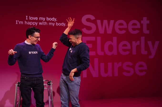 Dickie Hearts and Ryan J. Haddad stand on a pink stage with a pink wall behind them with the caption “I love my body. I’m happy with my body.” projected behind them in white.Ryan is a Lebanese American man with short black hair, brown eyes, round tortoiseshell glasses. Ryan wears a navy blue long-sleeve sweatshirt with the word “RYAN” printed in white across his chest and a white button-down shirt underneath it. His walker stands behind him. He wears blue jeans. Dickie Hearts has olive skin and dark chocolate-colored curly hair. He also wears a navy blue long-sleeve sweatshirt with the words “RYAN” printed in white across his chest. He wears light blue jeans. Ryan smiles widely and looks at Dickie. His arms are bent and he snaps his fingers. Dickie smiles with his mouth open and his eyes closed. He raises his right arm above his head and his arm is bent. His left arm is on his hip. He smiles at Ryan. Words  “Sweet Elderly Nurse” are to the right of Ryan and Dickie in large pink letters