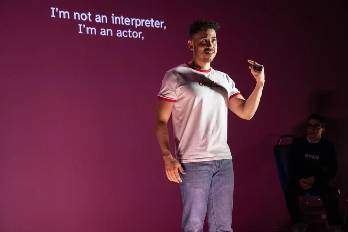 Dickie Hearts has olive skin and dark chocolate-colored curly hair. He wears a white tee shirt with a red neckline and red lined sleeves. The name “DICKIE” is written across his chest with two hearts dotting the I’s. He wears light blue jeans. Dickie smiles slightly, looking out to the audience and pointing to himself with his left hand and uses his index finger. He speaks in ASL. In the background, Ryan sits on the bus bench in the dark smiling at Dickie. Ryan is a Lebanese American man with short black hair, brown eyes, and round, tortoiseshell glasses. Ryan wears a navy blue long-sleeve sweatshirt with the word “RYAN” printed in white across his chest and a white button-down shirt underneath it. He wears blue jeans. His hands are clasped together.  