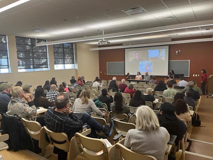 People looking at a presentation screen and panelists at the front of a classroom.
