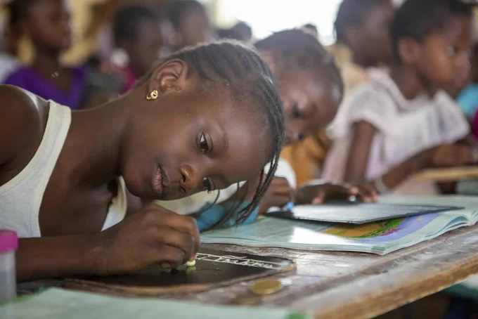 Young girl sitting at a desk and writing on a miniature chalkboard with a piece of chalk.