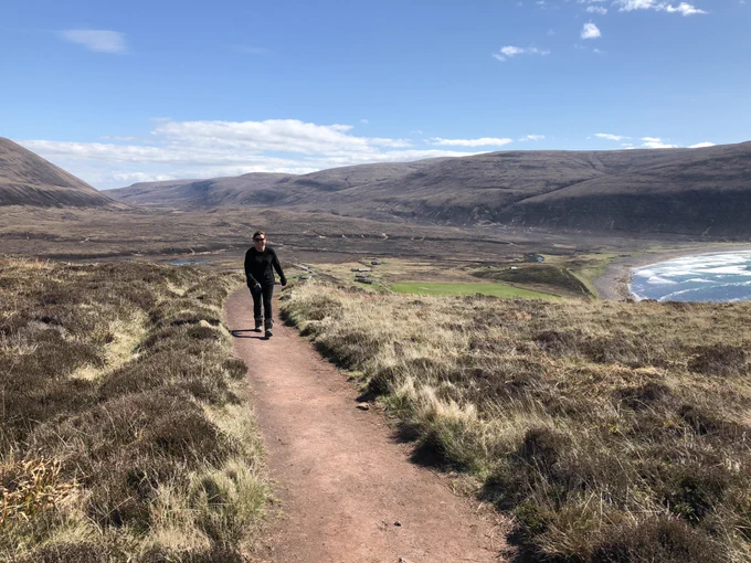 Lorraine Kelly walking on a path towards the Old Man of Hoy, with a view of heather-clad hills behind and Rackwick Bay to the right.