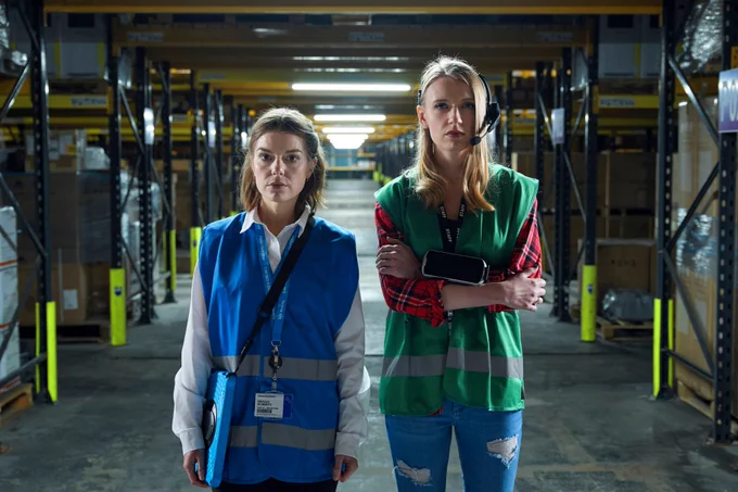 Image from Life and Death in the Warehouse showing Aimee-Ffion Edwards as Megan and Poppy Lee Friar as Alys. They are stood side by side looking directly at the camera with a warehouse behind them.