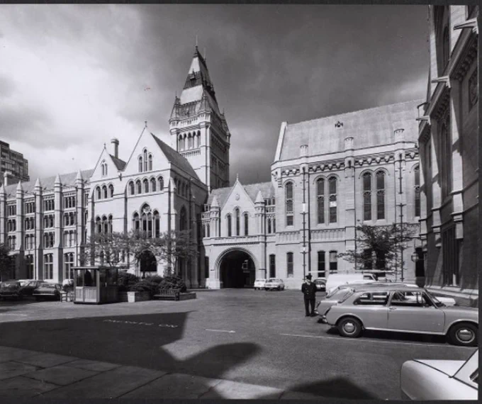 A black and white photo from 1972 in The Old Quad; shot at ground level from within the front quad angled towards the north east. It shows the museum, the arched front gatehouse leading out to Oxford Road, the Whitworth hall. There are cars in the foreground and a man in uniform.