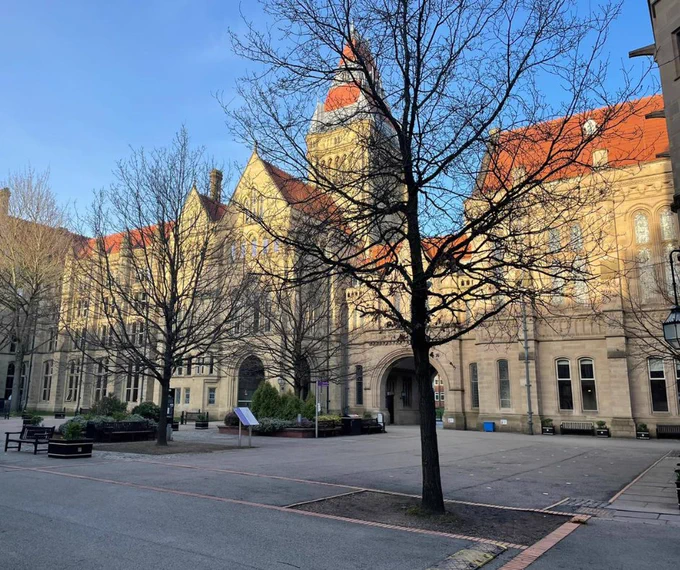 A bright colour photo of The Old Quad in 2023; shot at ground level from within the front quad angled towards the north east. It shows the museum, the arched front gatehouse leading out to Oxford Road, the Whitworth hall. There are trees in the foreground and a bright blue sky.