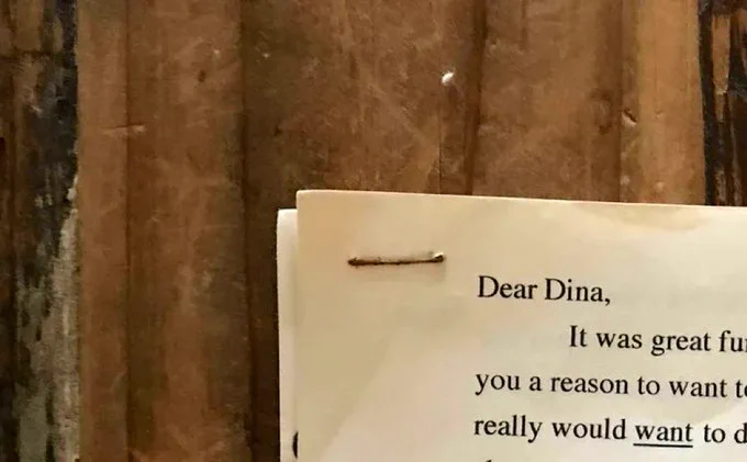 Picture of the corner of a typed, stapled page. The letter starts with "Dear Dina, it was great fun..." but most of the text is cut off. Background: the wood paneling of what appears to be a table.