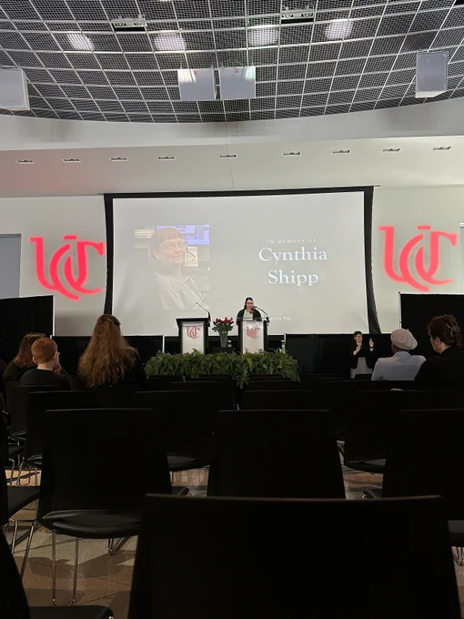 A projector screen reads "Cynthia Shipp" and shows a headshot of Cynthia. A person stands at a podium on a stage in front of the screen reading a tribute. Audience members sit in front of the stage listening.