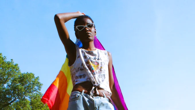 A person with sunglasses, holding a pride flag behind them