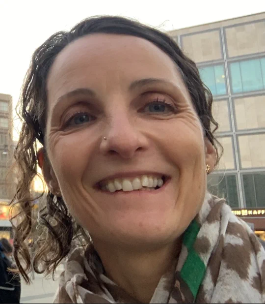 A headshot of Corinna, who is looking at the camera and smiling. She wears a scarf and has her brown, wavy hair tied back. She has a nose piercing.