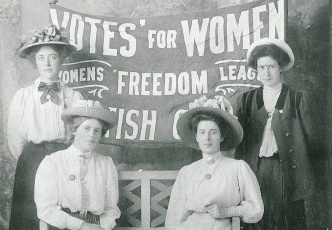 ID: Postcard photograph of four women in front of a Women’s Freedom League Votes for Women banner.