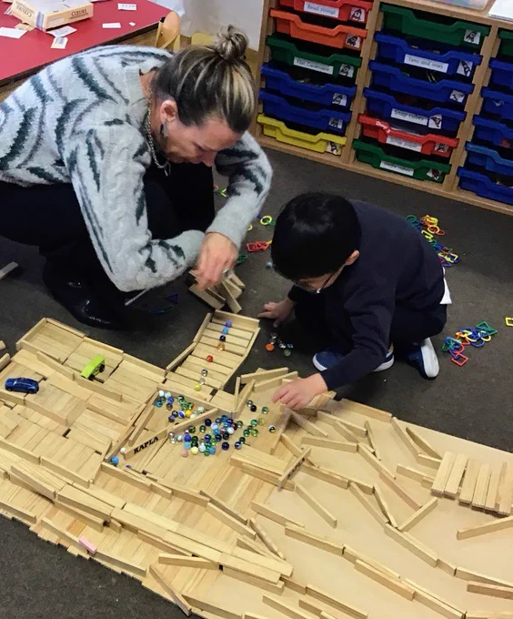 Kindergarten students have used a sloping board and Kapla to create a marble run for marbles. One student has made a line of marbles to represent the different planets that go around the sun, and is showing his teacher which is which.