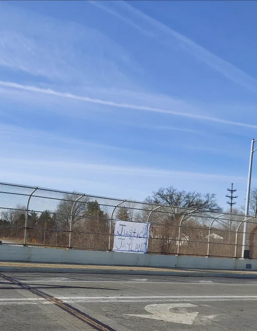 A white banner on a chain link fence. Banner reads "Justice 4 Jayland"