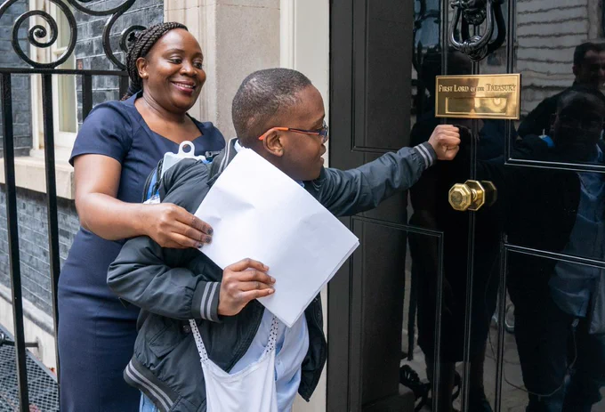 A boy and his mum hand in an open letter at No. 10 Downing Street. Mum is wearing a navy blue dress, while her son is wearing a navy bomber jacket and a blue shirt. 