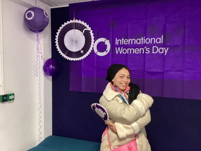 Photo of a student holding a sign with the International Women's Day logo and standing in the embrace equity pose, the flag behind them and lanterns and balloons.