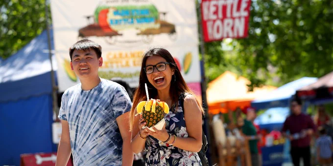 Two students walking and smiling at the camera, holding a drink in a pineapple. There are booths in the background for food and games. 
