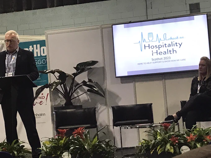 A white haired man in a suit stands behind a lectern to the left. In the centers is a screen which says ‘Hospitality Health’. To the right, a  blonde woman dressed in black is sitting down, listening