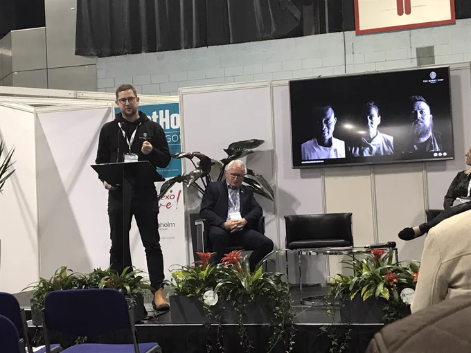 A man wearing glasses and dressed in black stands behinds a lectern and presents. Behind him a white haired man in a suit is sitting down, listening. On the wall is a screen showing a black and white photo of 3 chefs