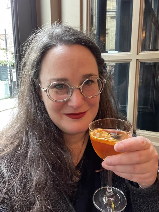 Helen, a white woman with long brown hair and red lippie sips an orange cocktail in a fancy coupe glass in a bar