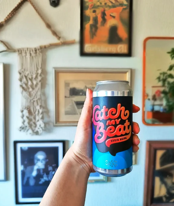 Hand holding up a can of beer in front of a wall full of artwork. 