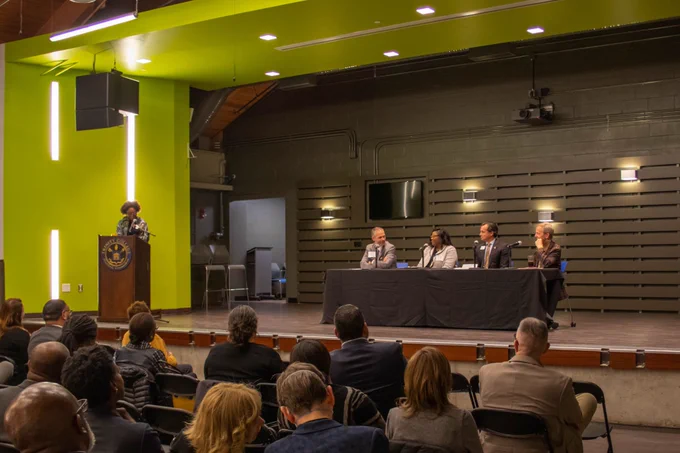 Four panelists sit at a table on stage, each behind a microphone. A moderator stands at a podium with a Trinity seal to the left. The stage is framed in a lit, lime green arch. Audience members sit in the foreground. 