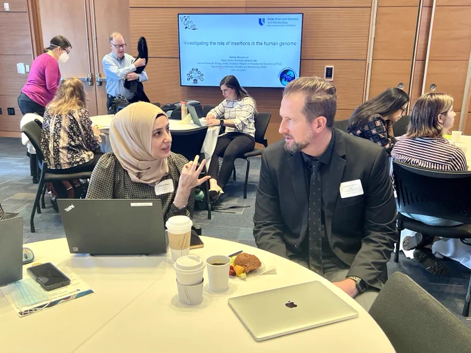 Attendees standing and sitting at tables in a conference room during a break at the Genomics Scientific Retreat. 
