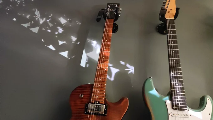 Two guitars hanging on the wall with tree leaf shadows across them.
